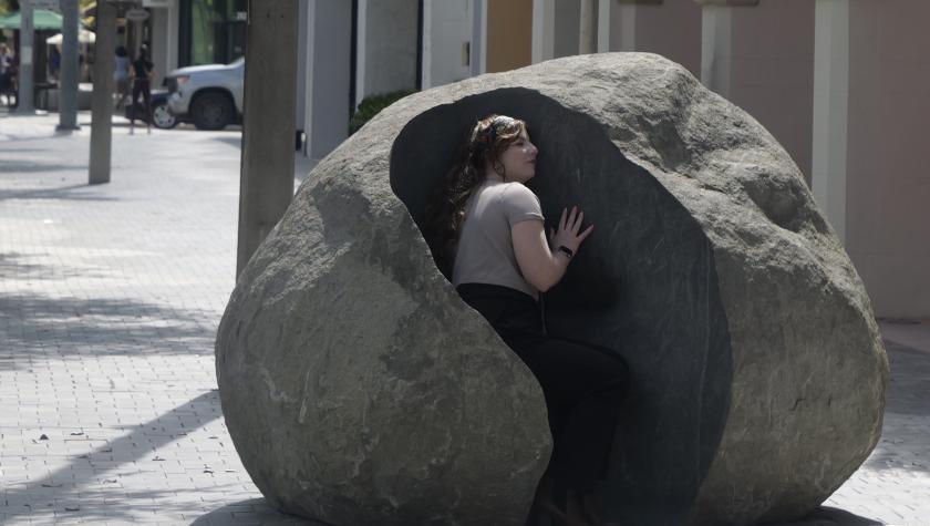 A woman sits inside a large rock sculpture with her ear pressed against it on a hot sunny day